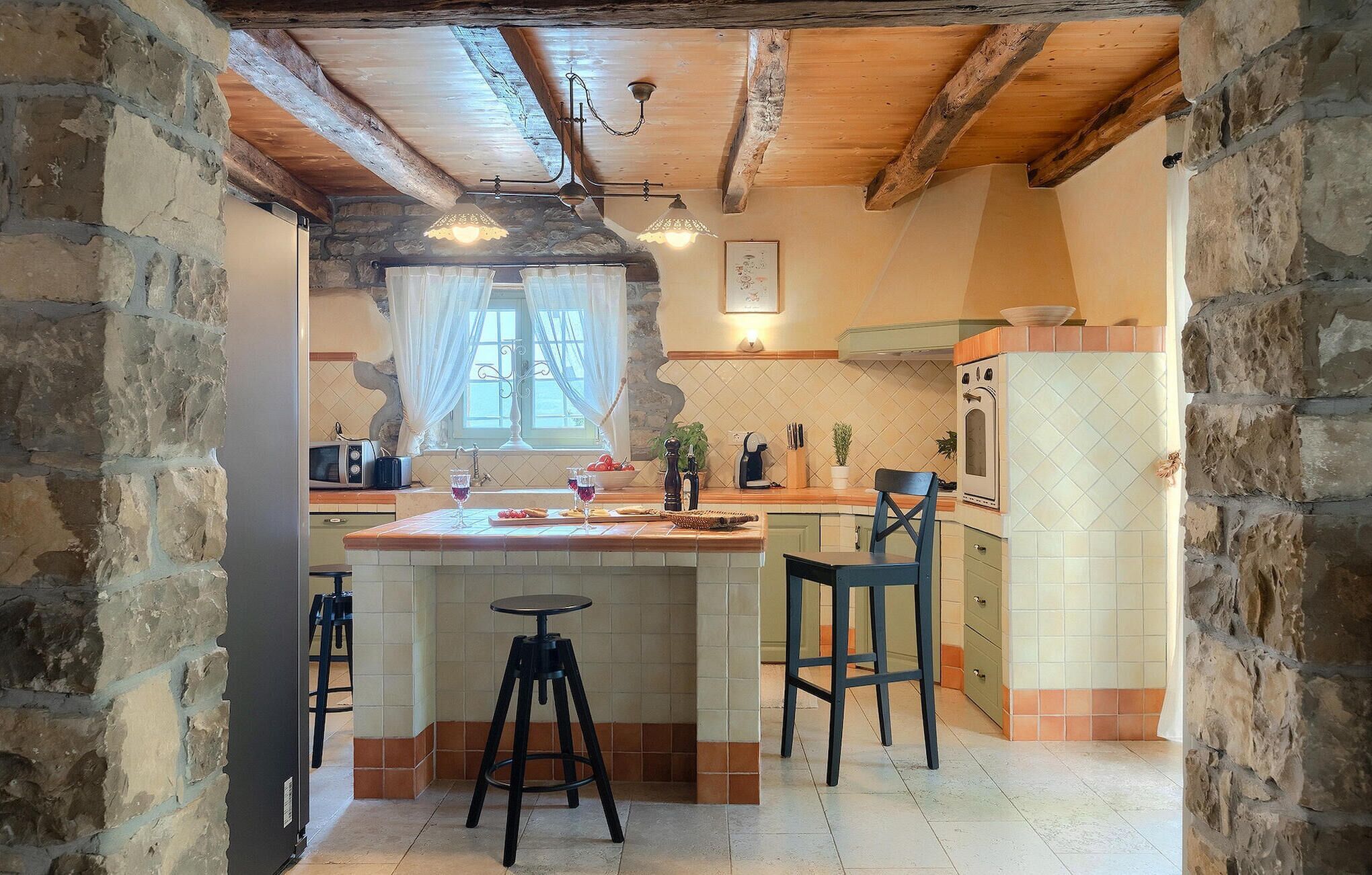A rustic kitchen with a stone wall, wooden beams, a tiled backsplash, and a central island with bar stools.