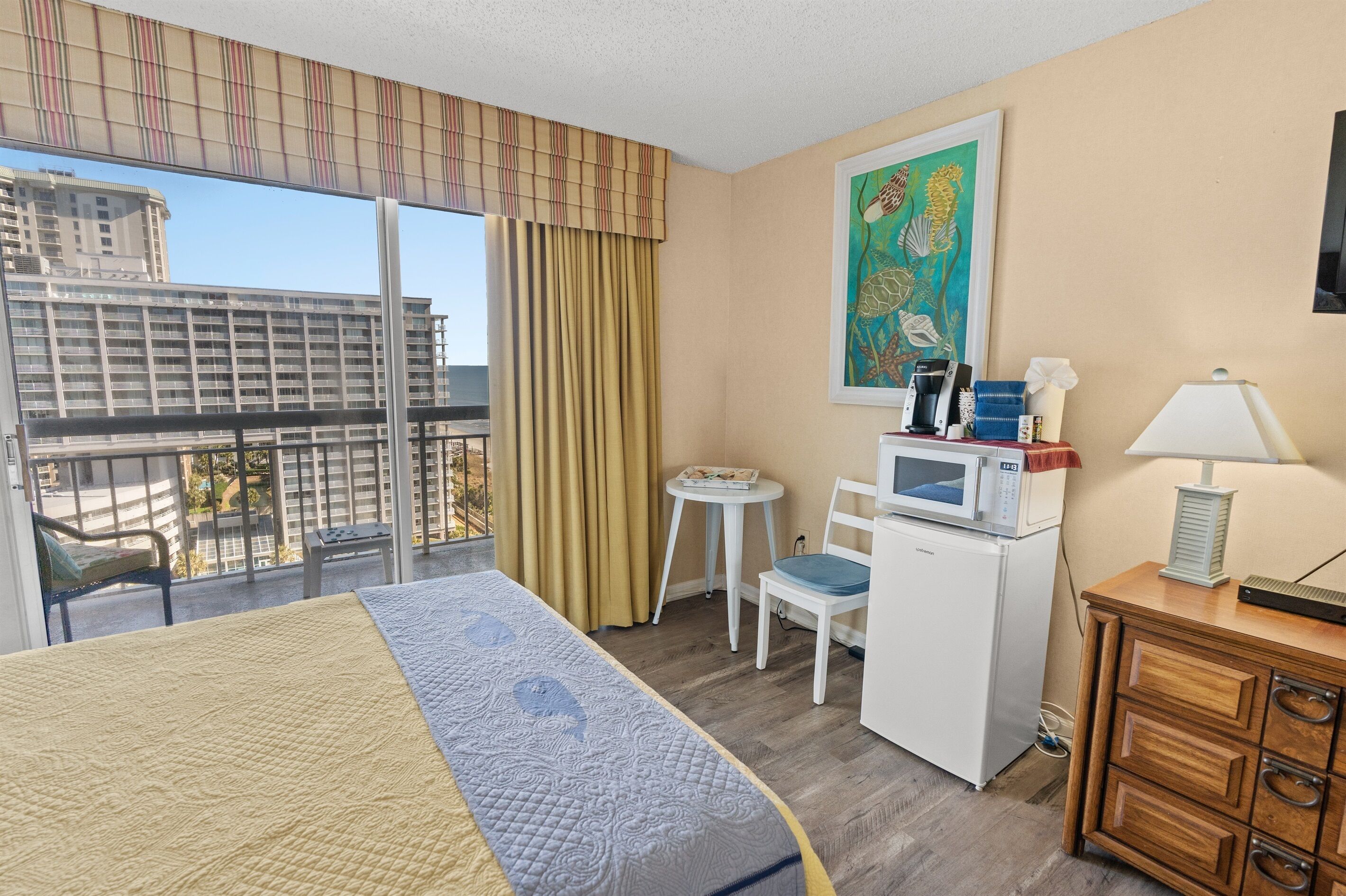 A hotel room with a bed, a desk with a microwave, a chair, a small table, a lamp, and a painting on the wall.