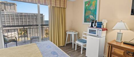 A hotel room with a bed, a desk with a microwave, a chair, a small table, a lamp, and a painting on the wall.