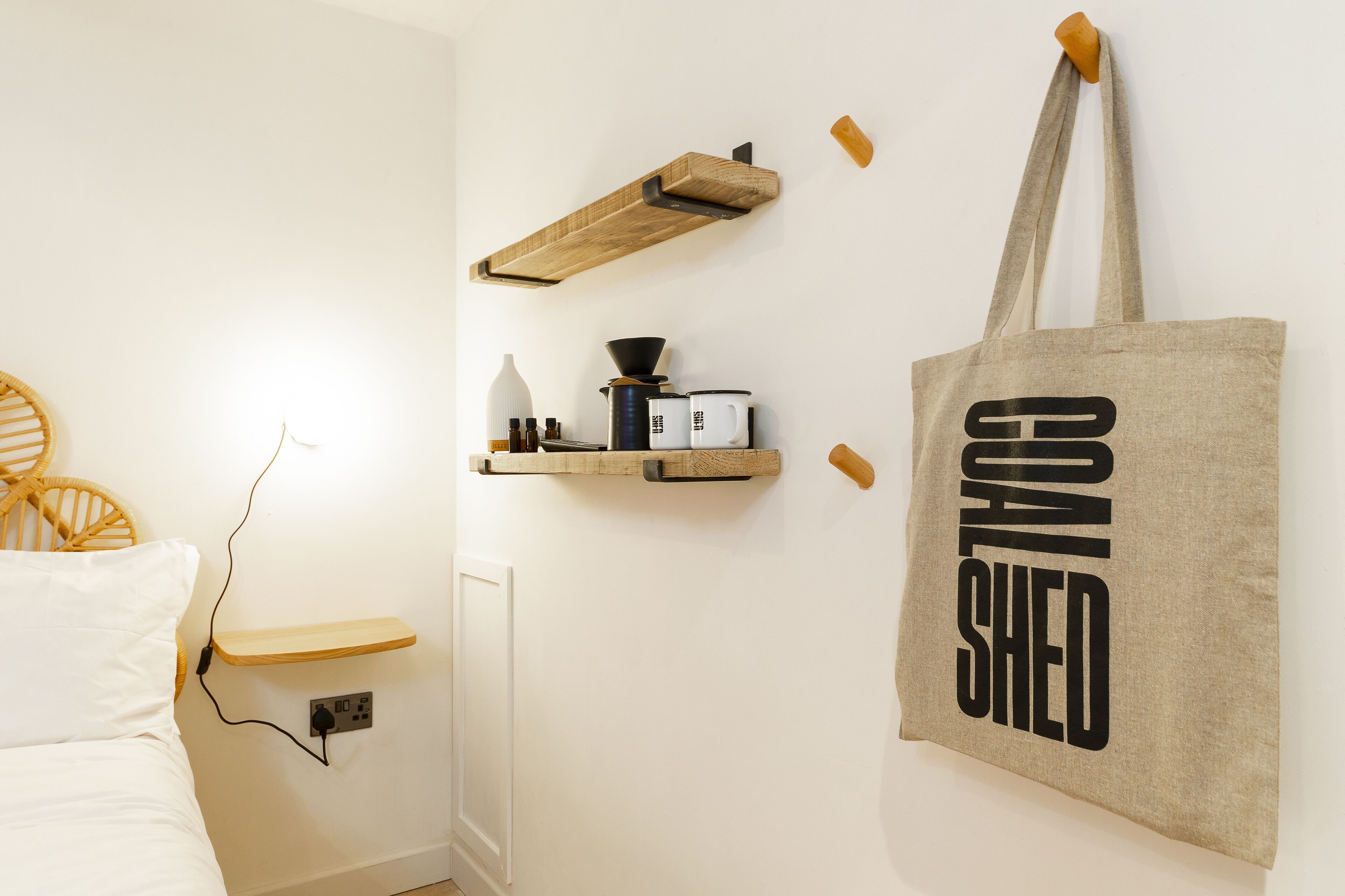 Un dormitorio con cama, una mesita de noche, un estante en la pared con una cafetera y una bolsa de tela con el texto 'COOK SHELD'.