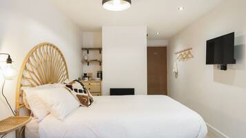 Un dormitorio con una cama de madera, una mesita de noche con lámpara, un televisor montado en la pared y una puerta que da a otra habitación.