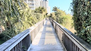 A wooden boardwalk with railings, surrounded by dense foliage and tall buildings in the background.