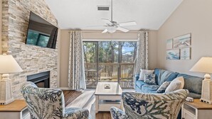 A living room with a stone fireplace, a ceiling fan, and a large window with curtains.