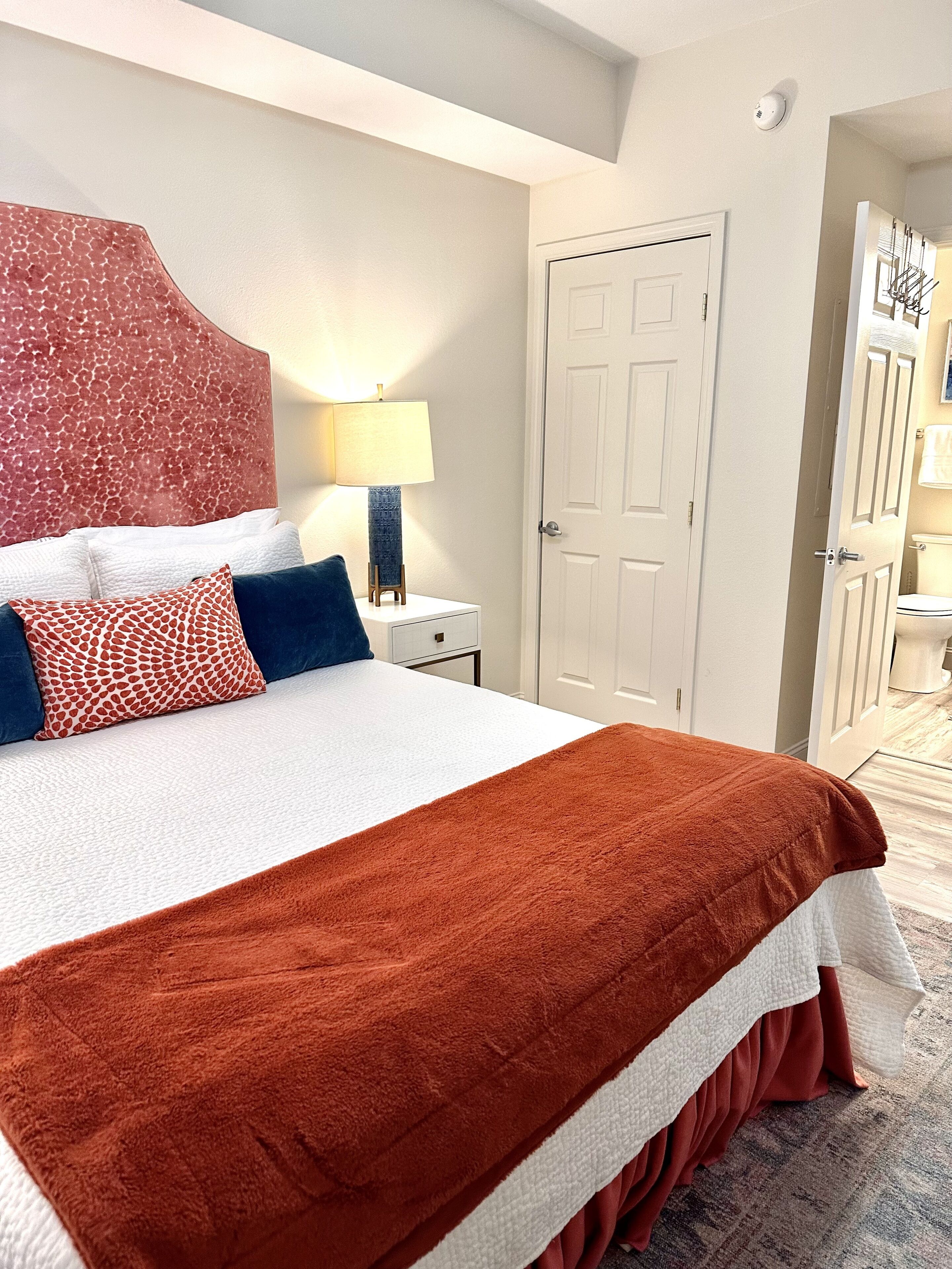 A neatly made bed with a red and white blanket, a patterned pillow, and a blue pillow. There is a bedside lamp, a white door, and a hallway leading to a bathroom.