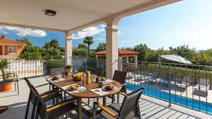 A covered outdoor dining area with a pool view, featuring a table set with food and drinks, surrounded by chairs. - Stunning home in Porec (Porec)