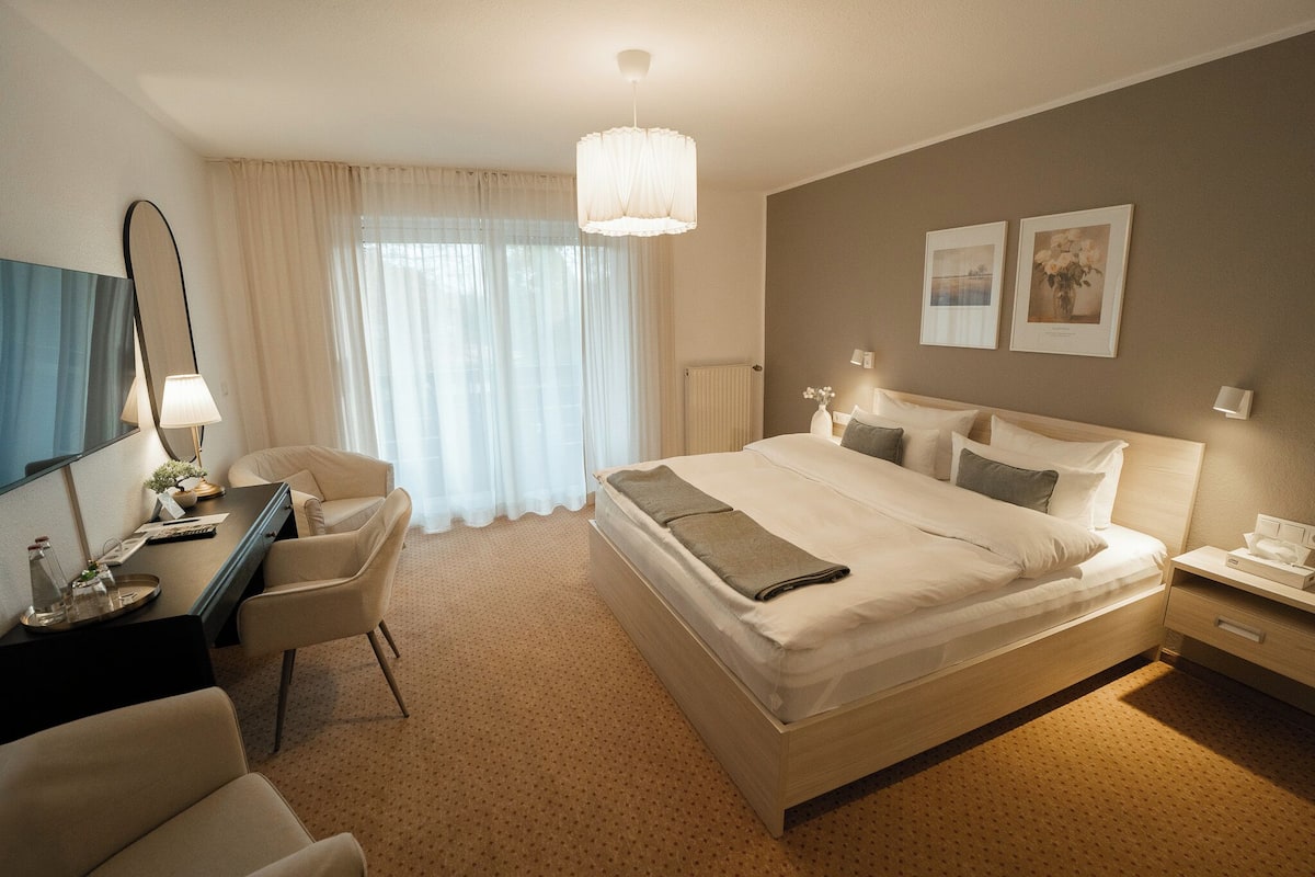 Ein Hotelzimmer mit einem großen Bett, einem Schreibtisch mit Stuhl, einem Nachttisch und einem großen Fenster mit Vorhängen.