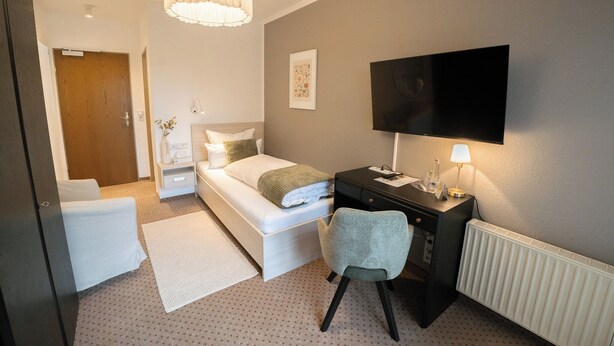 A bedroom with a bed, a desk, a chair, a TV, and a radiator. - The Resting Place (Lemgo)