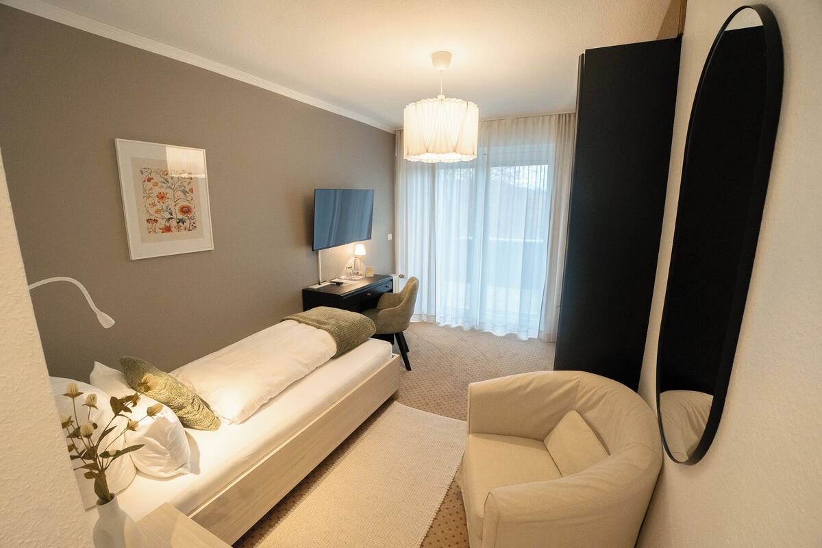 Ein modernes Hotelzimmer mit Bett, Schreibtisch, Stuhl, großem Spiegel und einem Fenster mit leichtem Vorhang.