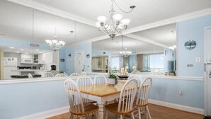 A dining room with a wooden table and white chairs, a chandelier, and a large mirror reflecting the kitchen.