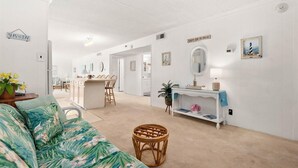 Un vaste espace de vie comprenant un canapé, un fauteuil en osier, un coin repas avec une table et des chaises, un coin cuisine et un couloir menant à une salle de bain.