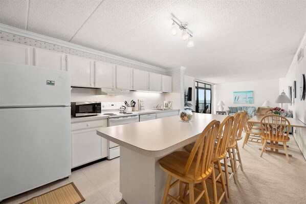 A modern kitchen with white cabinetry, a central island with chairs, and a dining area with a table and chairs. - Sea Terrace - Beautiful Ocean Views From this 2 Bedroom Condo with Outdoor Pool! (Ocean City)