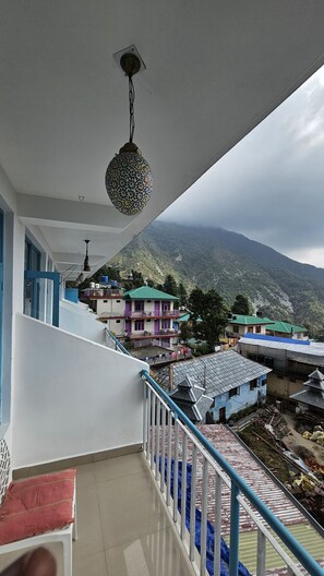 A balcony with a view of a mountainous landscape, a hanging lantern, and a staircase leading to a multi-story building with colorful facades. - LA SIESTA INDIA HOSTEL Mcleodganj (Dharamshala)
