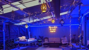 A cozy, dimly lit room with a neon sign stating "This is where the magic happens." There are multiple seating areas, a stage, and a projector on a table. - LA SIESTA INDIA HOSTEL Mcleodganj (Dharamshala)