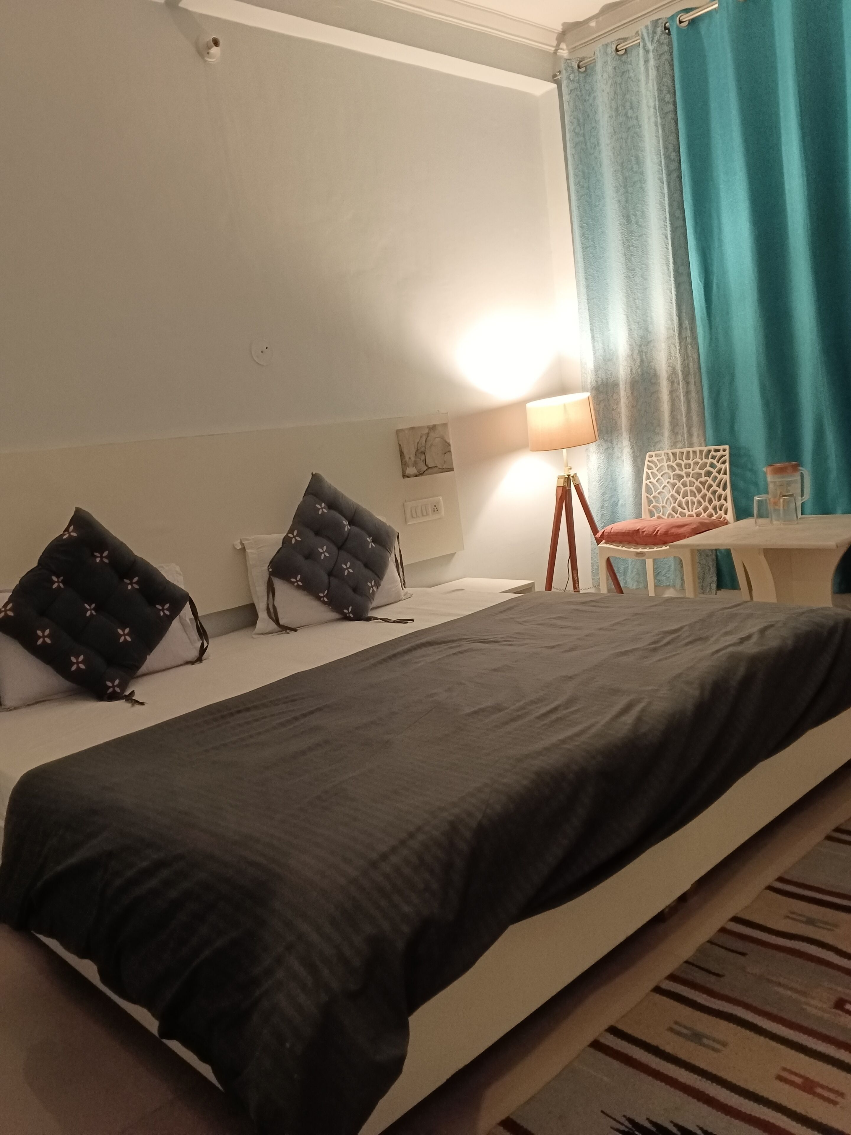A hotel room with a single bed, a small table with a lamp, a chair, and a window with curtains.