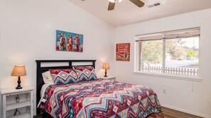 A bedroom with a bed, two bedside tables with lamps, a window with blinds, and a wall with a painting and a sign. - Affordable Rates!  The Grand Canyon getaway is a block from Downtown with A/C. (Williams)