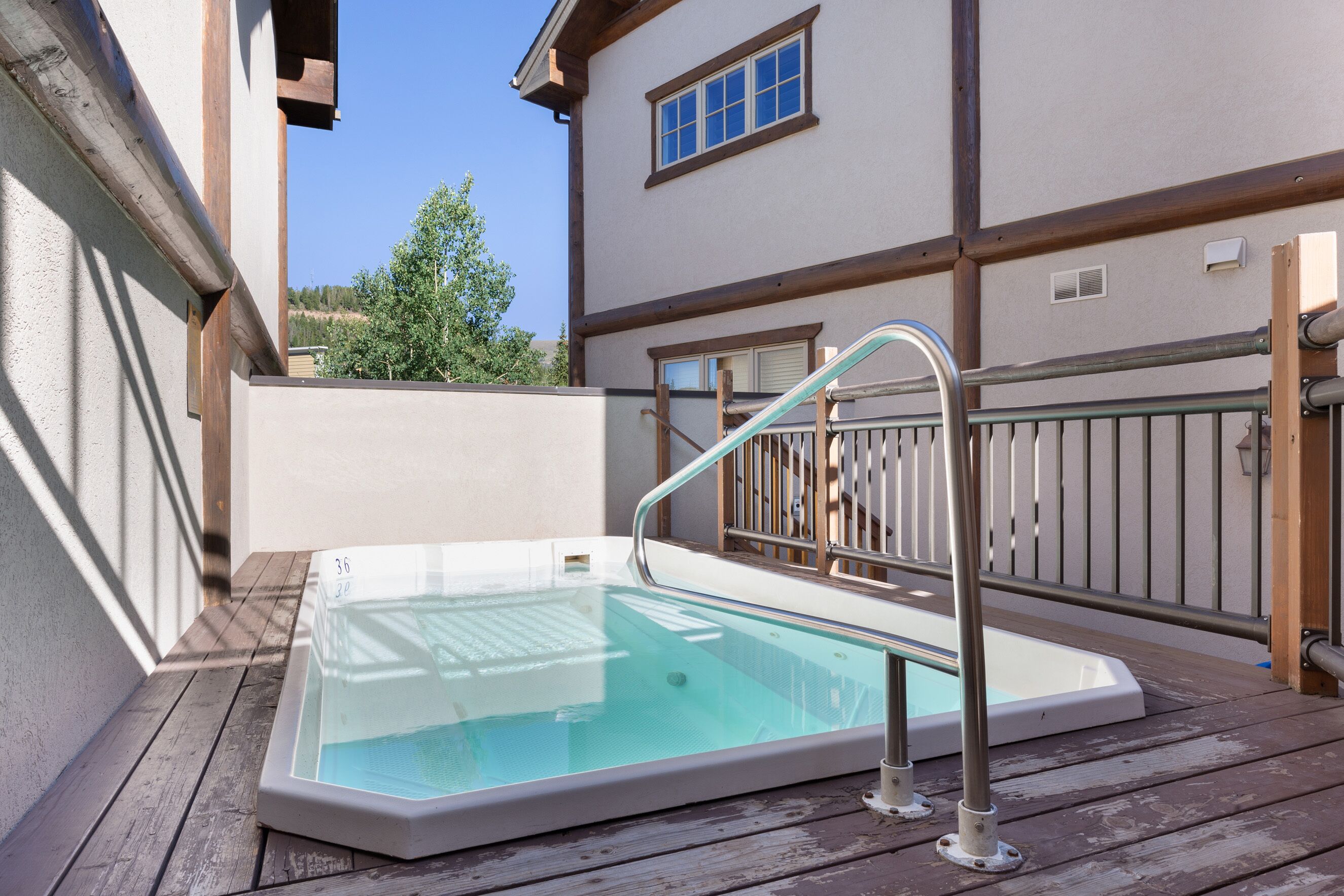 Une terrasse en bois avec un jacuzzi entouré de parois en verre, une rambarde en métal et un bâtiment doté d’une fenêtre et d’une porte.