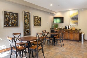 Uma área de refeições com mesas e cadeiras redondas, bancada de cozinha com pia e obras de arte nas paredes.