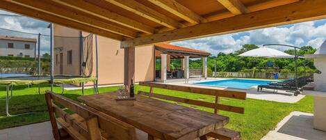 Un patio exterior con mesa y bancos de madera, una piscina y una zona de estar techada.