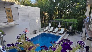 A swimming pool with lounge chairs and sun umbrellas, surrounded by potted flowers and a white wall with a window. - Apartments TP 777 (Budva)