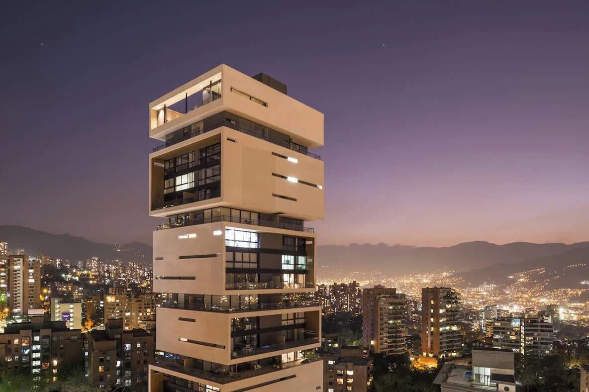 Un immeuble à plusieurs étages, doté de balcons et de grandes fenêtres, entouré d’un paysage urbain au crépuscule.