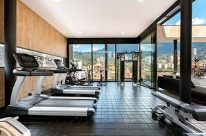 Une salle de sport moderne équipée de tapis de course, d’une machine de musculation et d’une zone avec une barre de traction, offrant une vue sur la ville grâce à de grandes fenêtres.