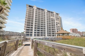 A multi-story hotel with balconies, a wooden boardwalk, and a beach area with a thatched structure.