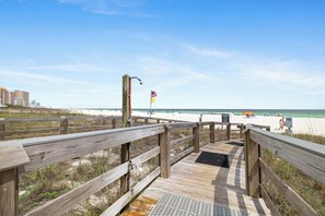 A wooden boardwalk leading to a beach with a flag, a sandy area, and buildings in the background.