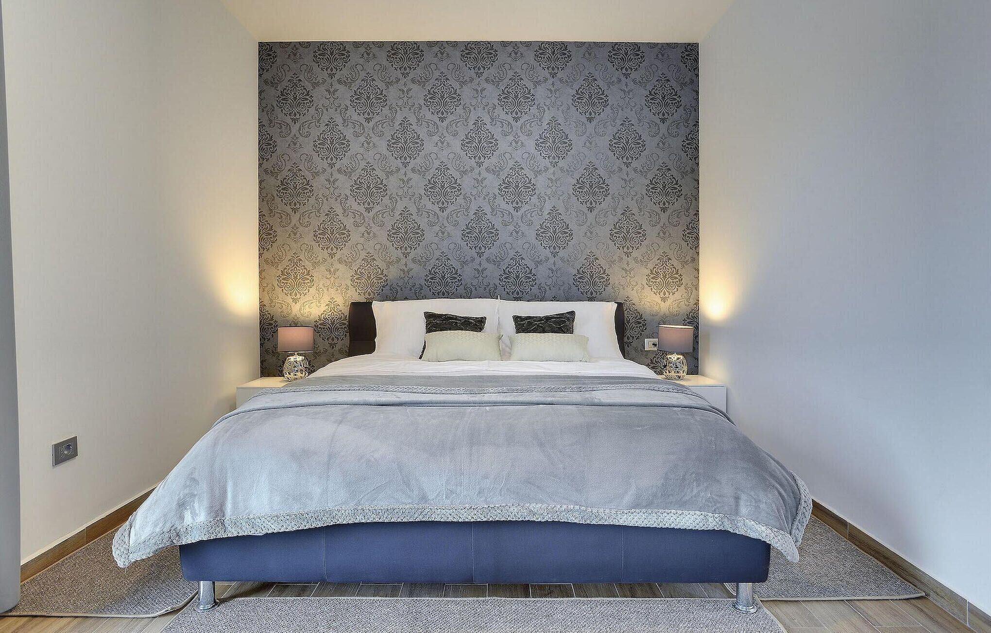 A narrow room with a bed, two bedside lamps, and a patterned wallpaper.