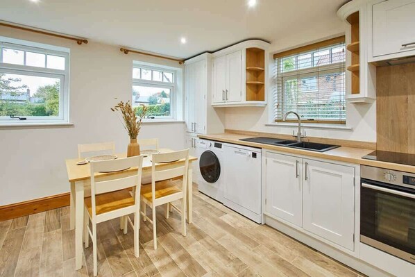 A modern kitchen with white cabinetry, a built-in washing machine, a sink, and a stove. There is a dining table with chairs and a vase with dried plants.