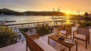A terrace with wooden furniture, a table with a bottle and glasses, and a view of a body of water during sunset. - Beach front home in Preko (Preko)