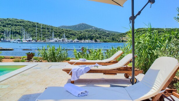 A poolside area with lounge chairs and an umbrella, overlooking a marina with boats and a mountainous backdrop. - Beach front home in Preko (Preko)