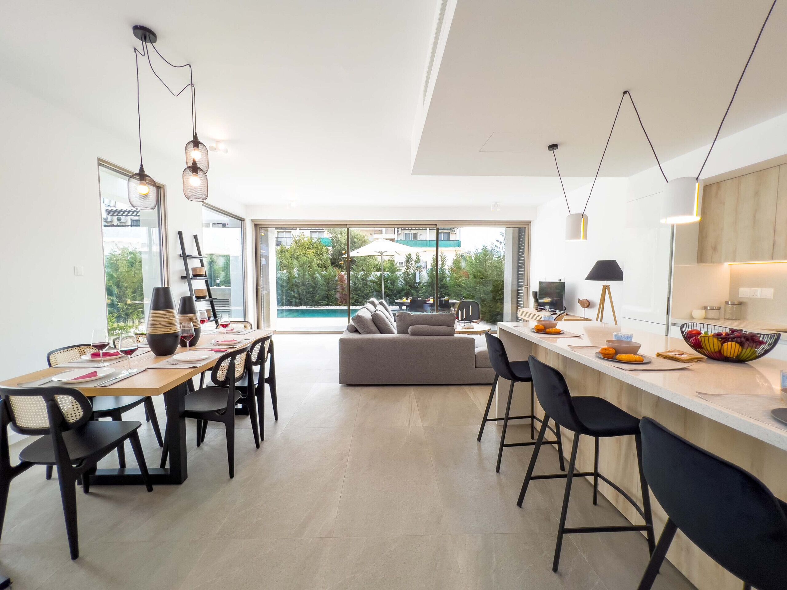 A modern kitchen with a dining area, a large window with a view of greenery, and a pool visible through the glass doors.