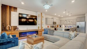 A modern living room with a flat-screen TV, a fireplace, and a dining area with a kitchen in the background.