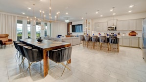 A modern kitchen with white cabinets, a dining area with a large wooden table and chairs, and a TV mounted above the counter.