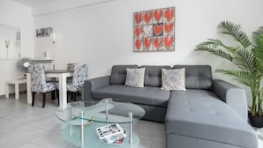 Un salon moderne avec un canapé d’angle gris, une table basse en verre et un coin repas avec des chaises à motifs.