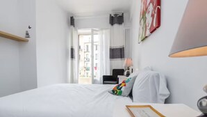 Une chambre d’hôtel avec un lit, une table de chevet avec une lampe, une petite étagère et une vue sur la rue par une porte ouverte.