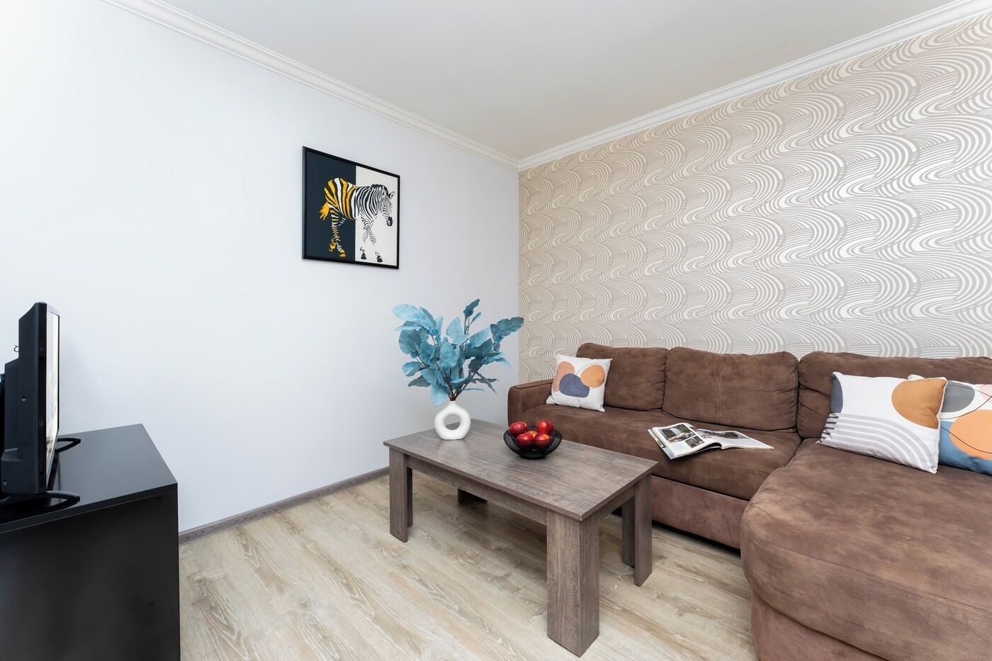 A living room with a brown sofa, a coffee table with a vase and apples, a framed picture on the wall, and a patterned wallpaper.
