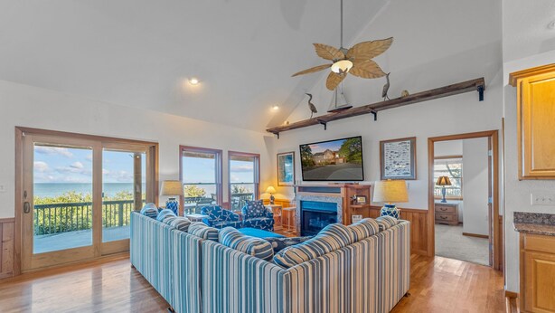 A spacious living room with a large striped sofa, a fireplace, a television, and a balcony with a view of the ocean. - Soundfront Dog Friendly w/ Hot Tub, Elevator, Private Sound Access, Media Room (Buxton)