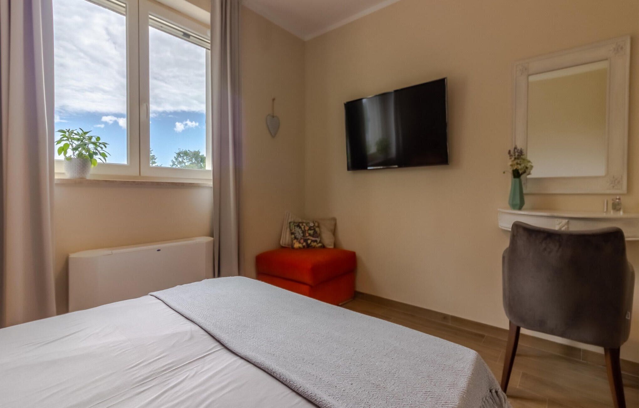 A hotel room with a bed, a red chair, a TV, a window with a plant, and a mirror.