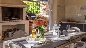An outdoor dining area with a stone fireplace, a grill, and a table set for two with a bottle of wine, a vase of flowers, and a candle.