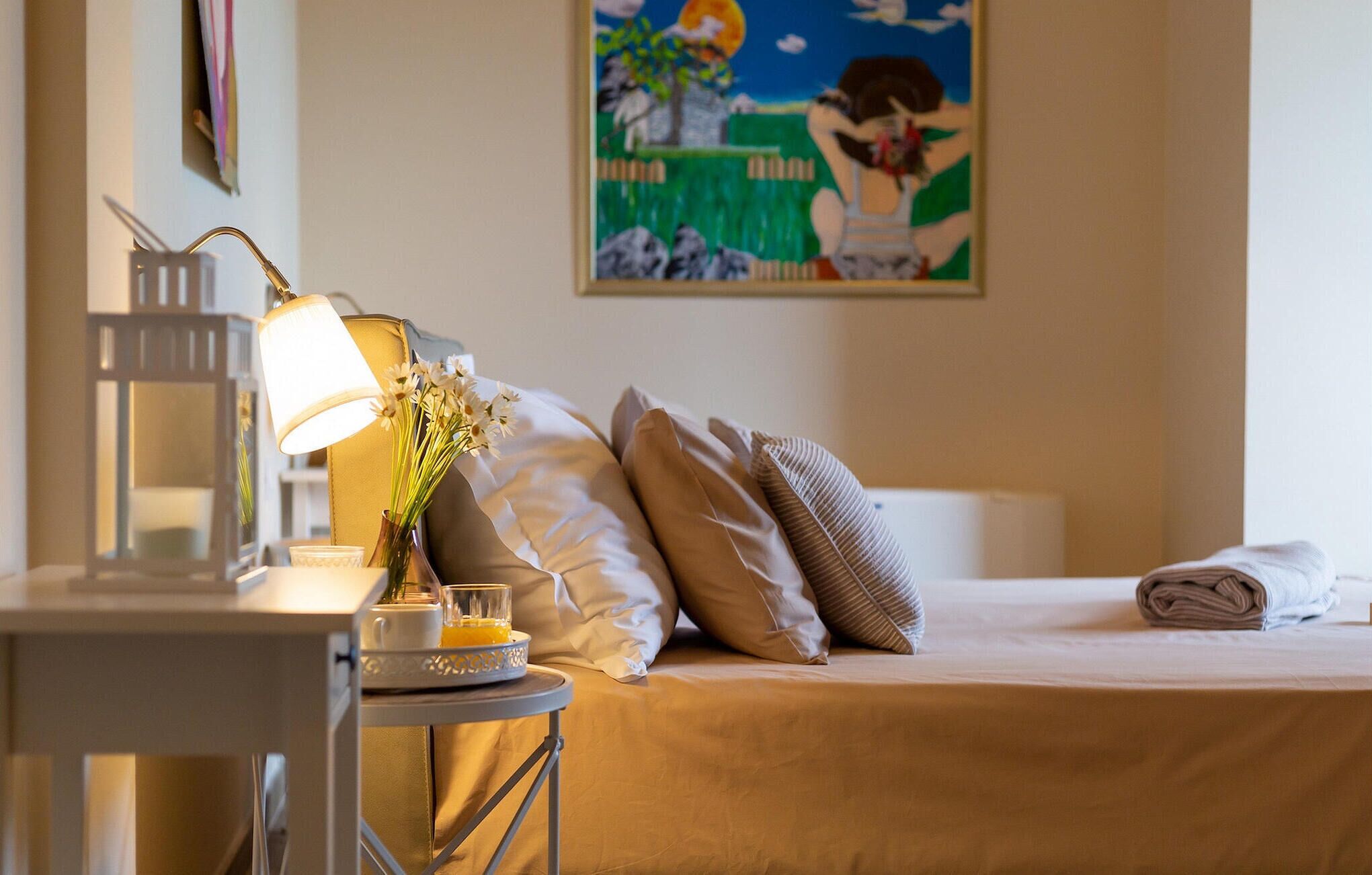 Ein Hotelzimmer mit Bett, Kissen, Nachttisch mit Lampe, Blumen in einer Vase und einem Bild an der Wand.