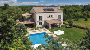 A two-story house with a swimming pool, a covered patio, and a view of the surrounding greenery and mountains.