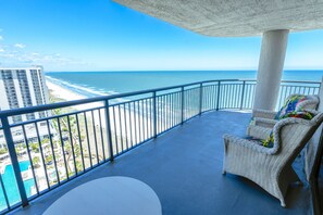 Un balcón con vista al océano y la playa, una silla de mimbre y techo de concreto.