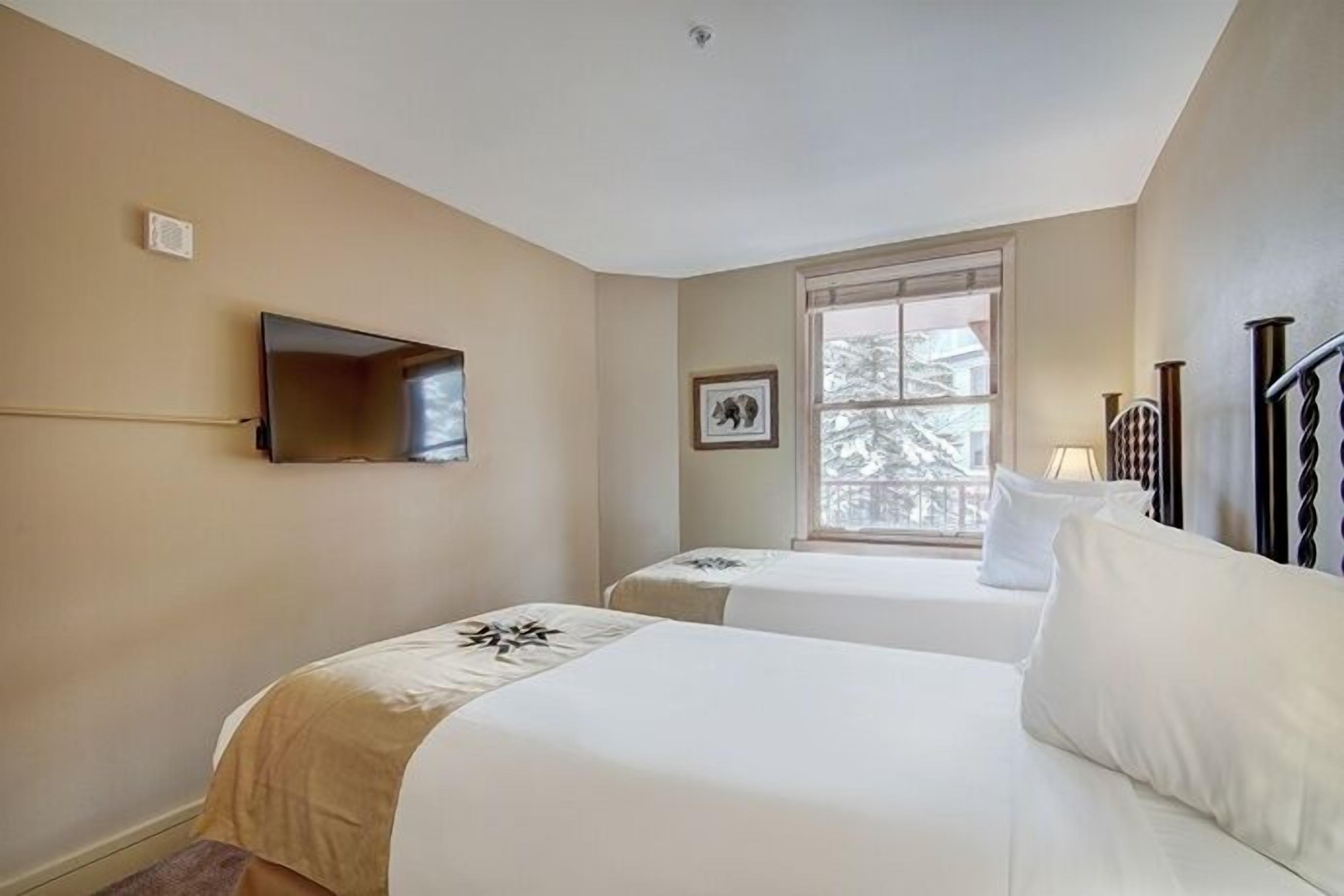 Une chambre d’hôtel avec deux lits, un téléviseur fixé au mur, une fenêtre donnant sur des arbres et un cadre photo accroché au mur.