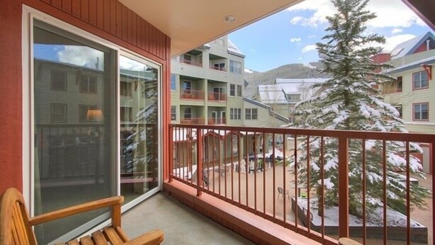 Un balcon avec un banc en bois, une porte vitrée et une vue sur un arbre et des bâtiments recouverts de neige.