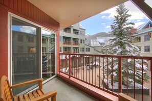 Un balcon avec un banc en bois, une porte vitrée et une vue sur un arbre et des bâtiments recouverts de neige.