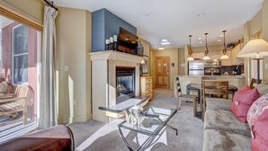 Un salon avec une cheminée, une table basse en verre, un canapé et, au fond, un coin cuisine.