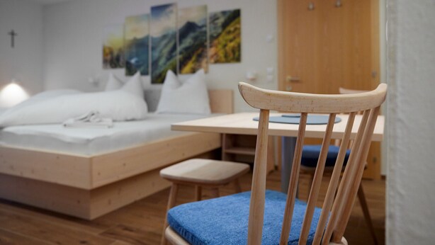 A bedroom with a bed, a desk, and a chair. There are paintings on the wall and a wooden door in the background. - 5003 Alice im Alpenhaus Montafon (Gargellen)