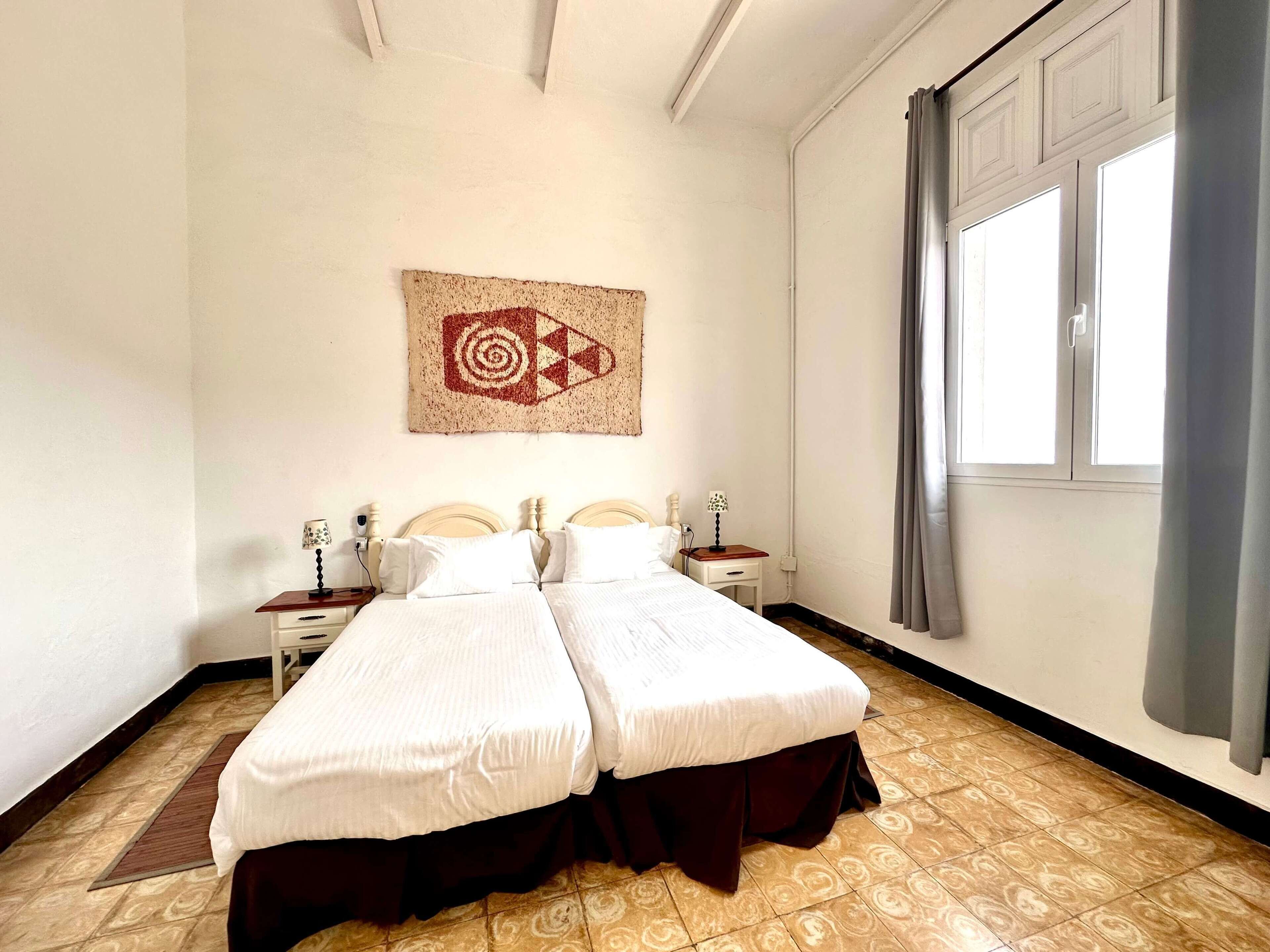 A double bed with two nightstands, a wall-mounted artwork, a window with curtains, and patterned flooring.