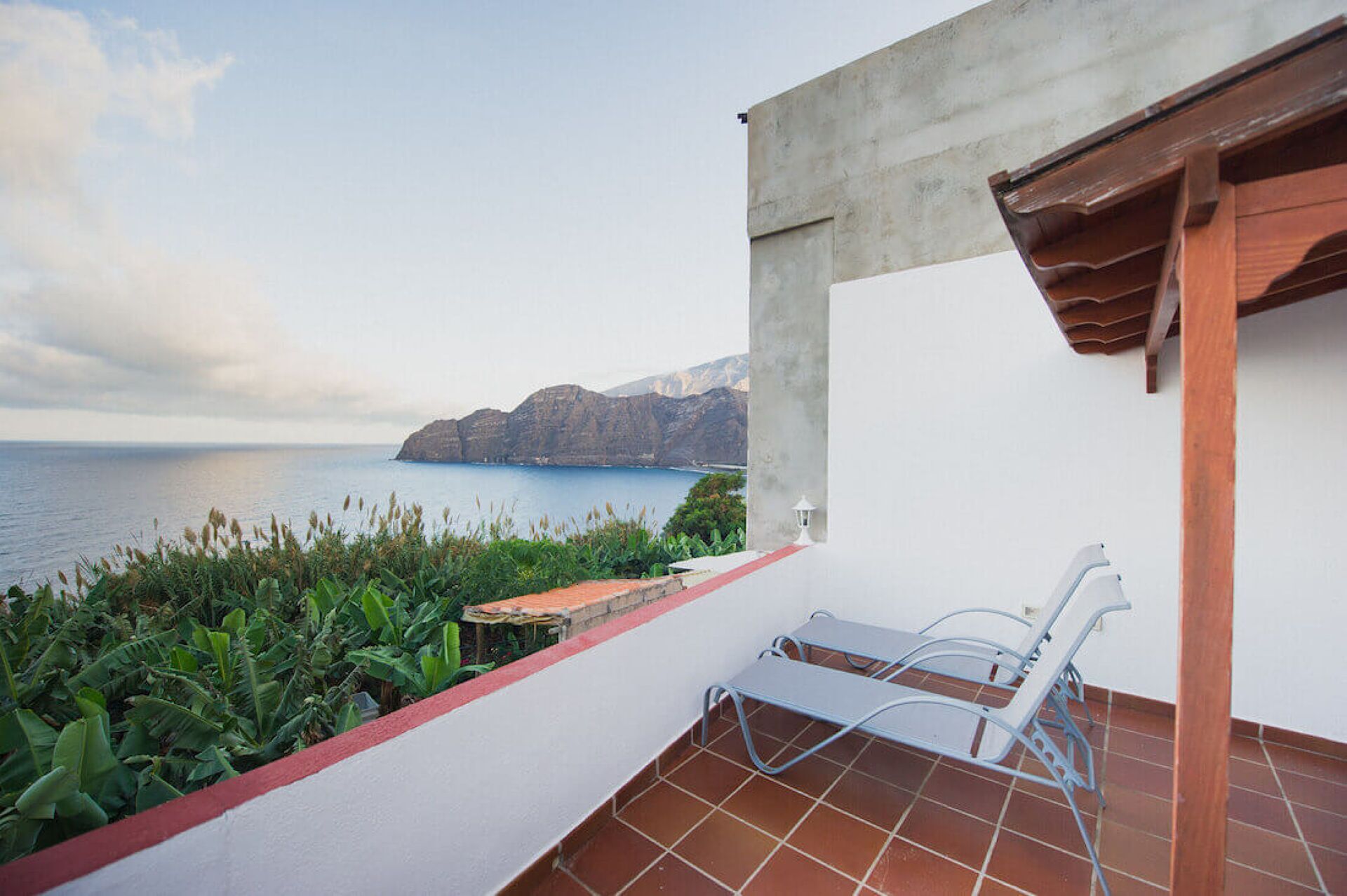 Un balcón con vistas al mar y a las montañas, una pared blanca y una silla de metal.